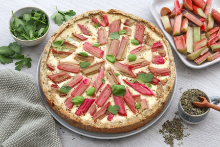 Ein Rhabarberkuchen auf einem Tisch, neben Kräutern und geschnittenem Rhabarber.