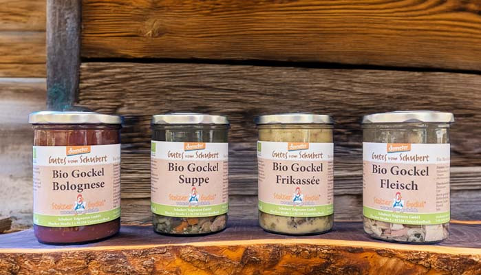 Vier Gläser mit verschiedenen Bio-Gockel-Produkten vor Holzhintergrund. Text: Bio Gockel Bolognese, Suppe, Frikassee, Fleisch.