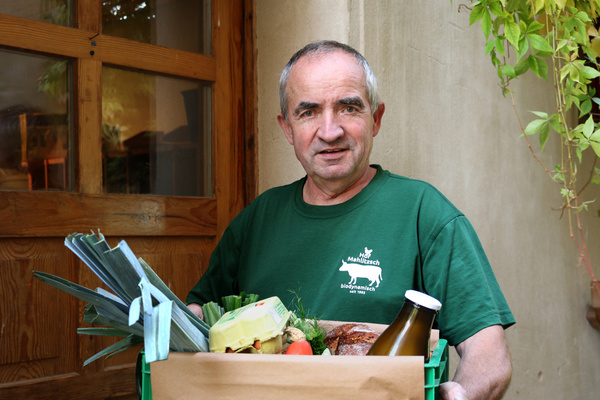Unser Lieferfahrer Andrea steht vor einer Haustür und hält eine Kiste mit verschiedenen frischen Lebensmitteln, darunter Gemüse, Eier und eine Flasche Milch.