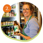 Karoline wählt gerade eine Flasche frische Mahlitzscher Vollmilch für einen Kunden aus dem Kühlschrank. Sie lächelt in die Kamera. Im Hintergrund sind weitere Milchprodukte zu sehen.