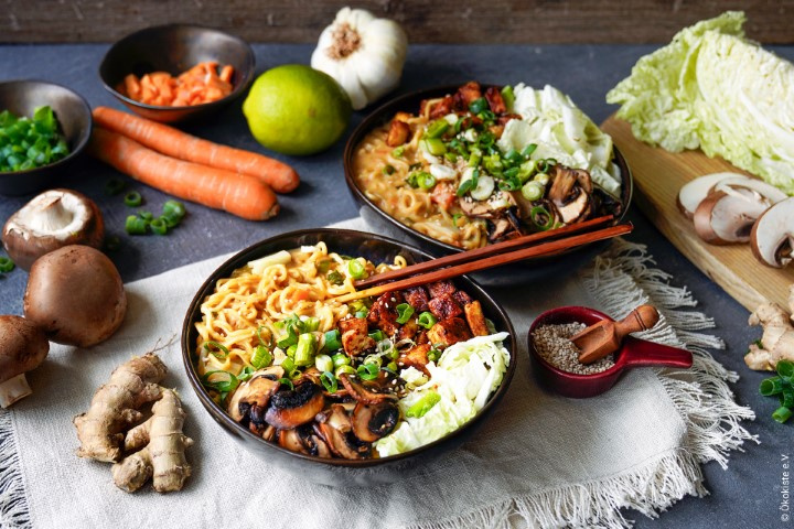 Das Bild zeigt zwei Schalen mit Ramen, die mit frischen Zutaten wie Pilzen, Frühlingszwiebeln und Tofu angerichtet sind. Rund um die Schalen liegen verschiedene frische Zutaten wie Karotten, Ingwer und Knoblauch.