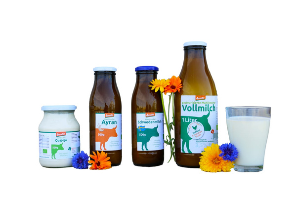 Das Bild zeigt verschiedene Molkereiprodukte in Glasflaschen und einem Glas, dekoriert mit Blumen. Die Produkte sind von der Marke Demeter und umfassen Quark, Ayran, Schwedenmilch und Vollmilch.