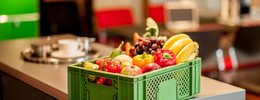 KI generiert: Das Bild zeigt eine grüne Kiste mit verschiedenen Obst- und Gemüsesorten auf einem Tisch in einem Innenraum. Im Hintergrund sind unscharf einige Stühle und Tische zu sehen.
