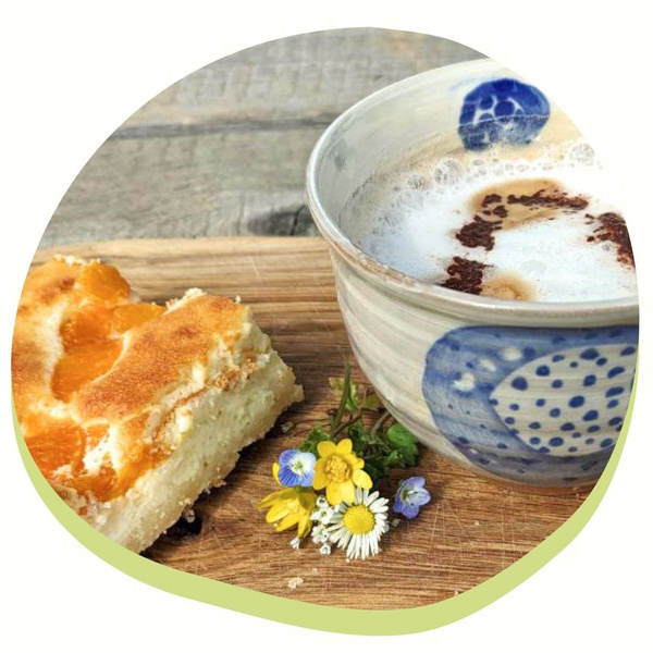 KI generiert: Das Bild zeigt eine Tasse Cappuccino oder Milchkaffee neben einem Stück Kuchen mit einem Fruchtbelag, auf einem Holzbrett mit einigen kleinen Blumen. Der Hauptinhalt ist eine ansprechende Kaffeepause oder ein Snack.