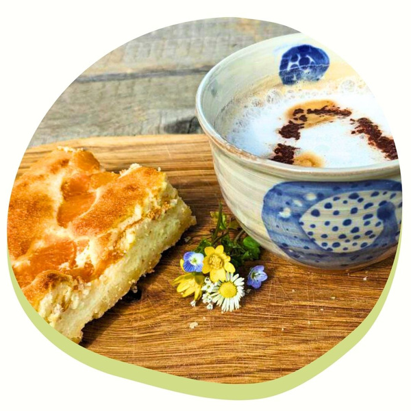 KI generiert: Ein Stück Kuchen, eine Tasse Cappuccino und Blumen auf einem Holzbrett.