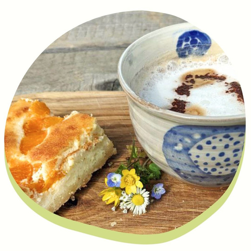 KI generiert: Das Bild zeigt eine Tasse Cappuccino oder Milchkaffee neben einem Stück Kuchen mit einem Fruchtbelag, auf einem Holzbrett mit einigen kleinen Blumen. Der Hauptinhalt ist eine ansprechende Kaffeepause oder ein Snack.